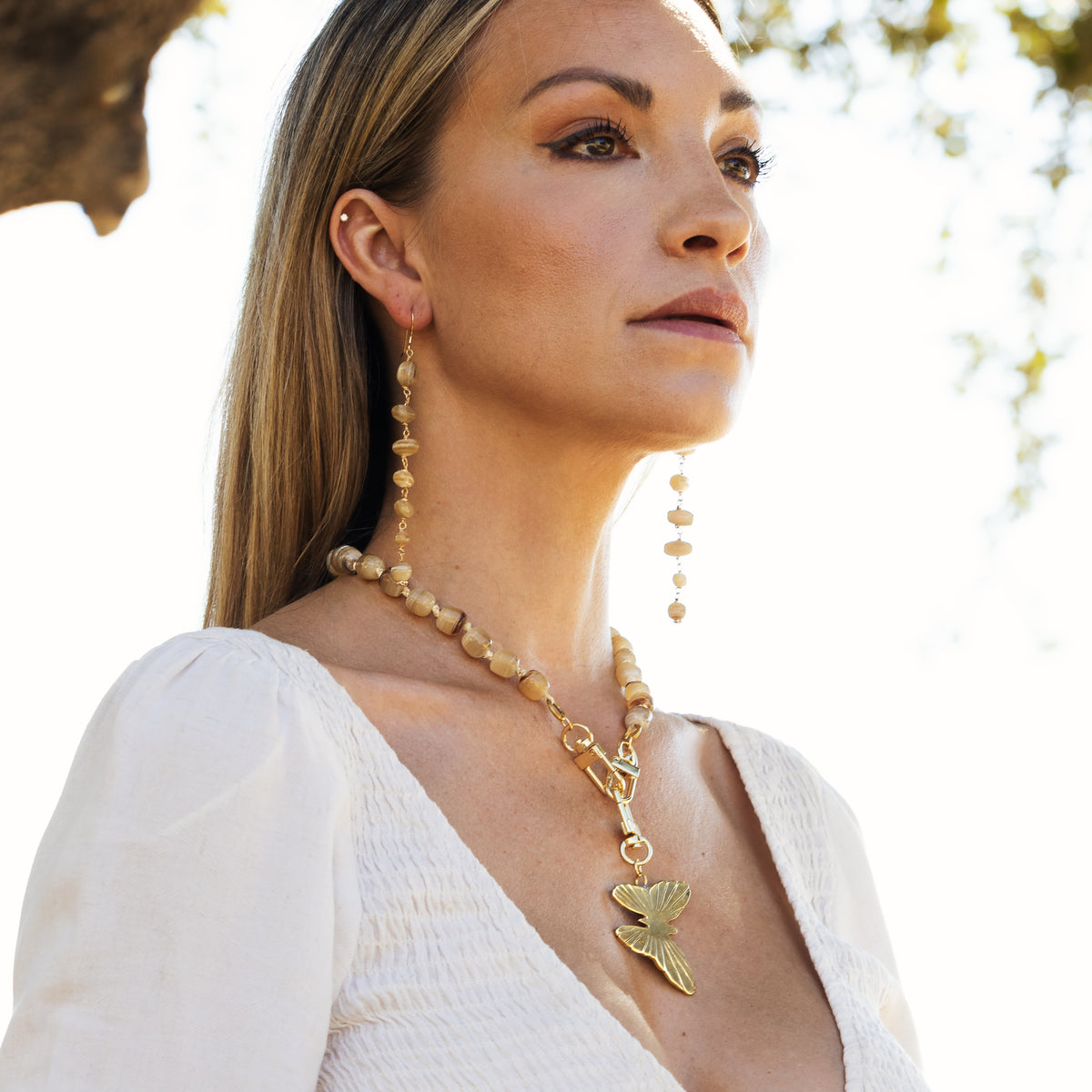 A woman wearing a gold necklace with a bird pendant and matching drop earrings, styled with a white top against a bright outdoor background.